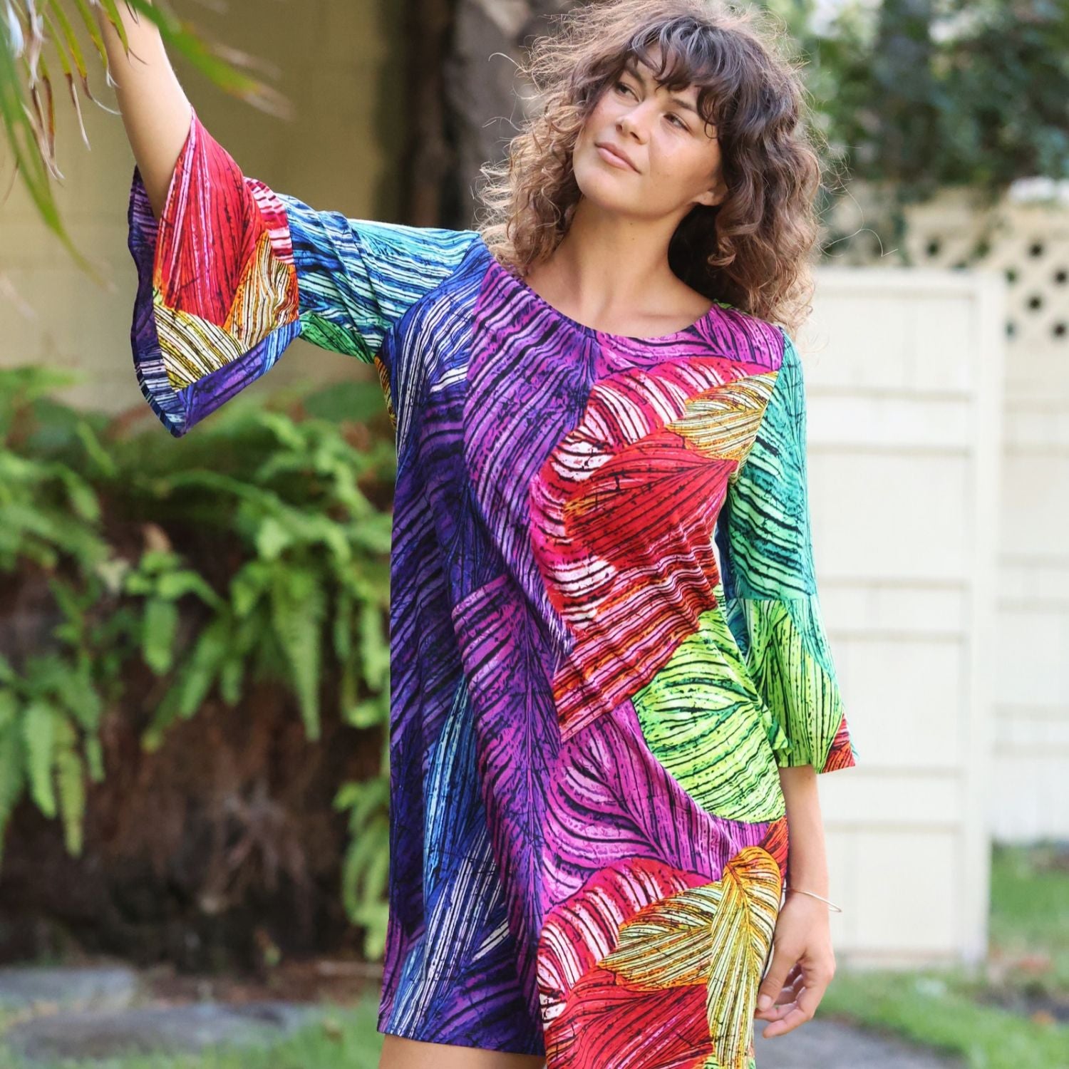 A woman wearing a vibrant, multicolored leaf-patterned tunic dress with three-quarter sleeves, standing outdoors in front of a white fence and green plants.