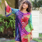 A woman wearing a vibrant, multicolored leaf-patterned tunic dress with three-quarter sleeves, standing outdoors in front of a white fence and green plants.
