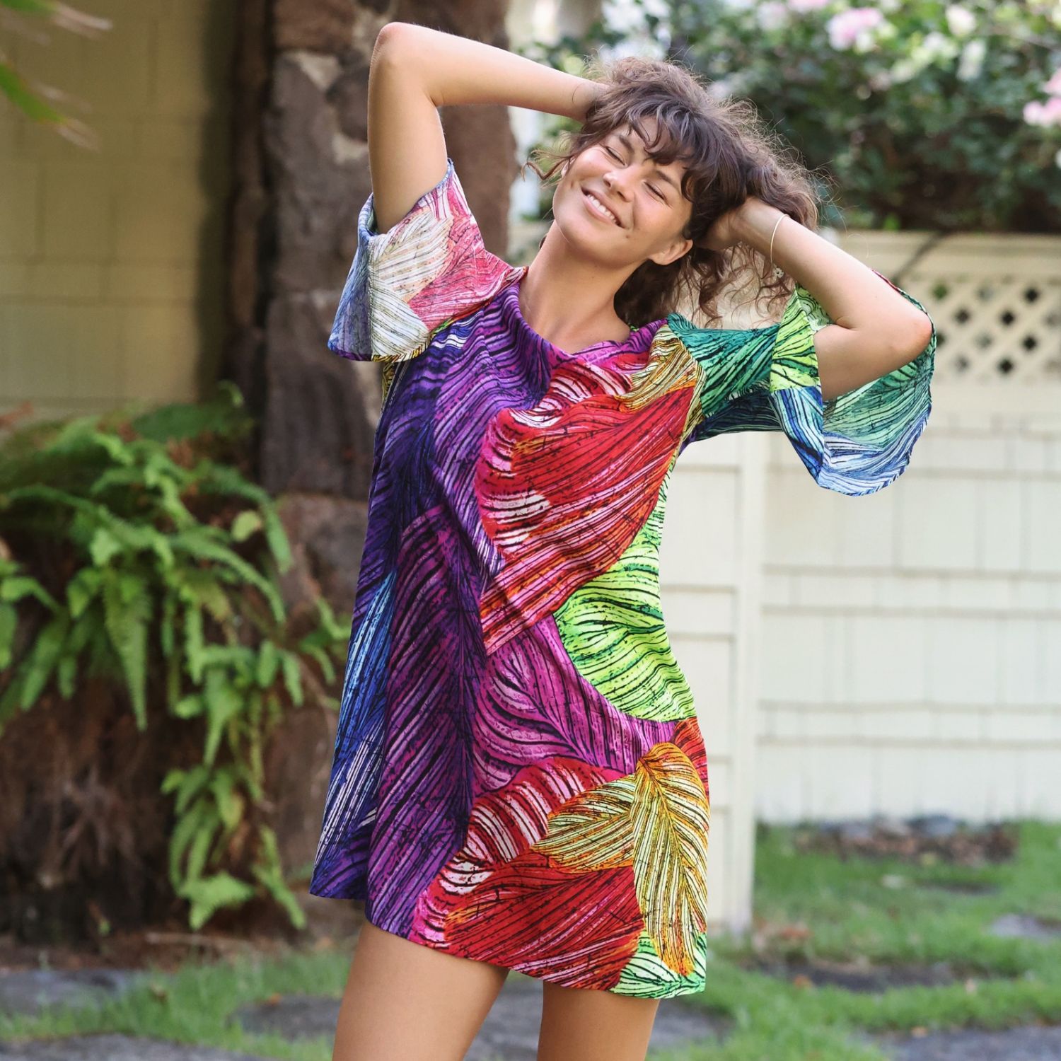 A woman wearing a colorful tropical leaf print mini dress, posing outdoors with her hands behind her head in a garden setting.