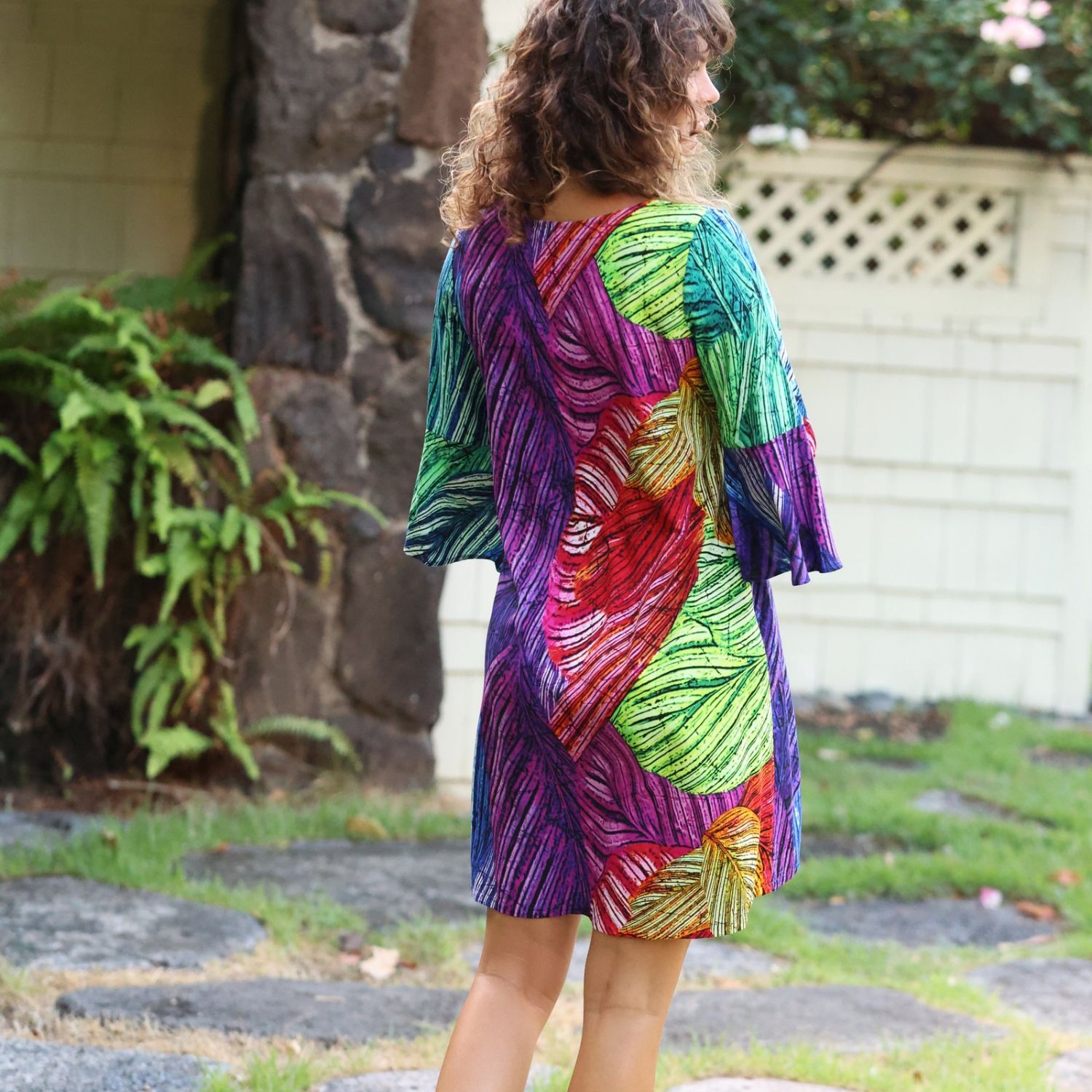 A woman wearing a vibrant, multicolored leaf-patterned dress, photographed from behind in an outdoor backyard setting with a stone pillar and white fence in the background.