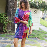 A woman wearing a vibrant, multicolored tropical print dress with three-quarter bell sleeves, standing outdoors on a stone path with greenery and a white shed in the background.