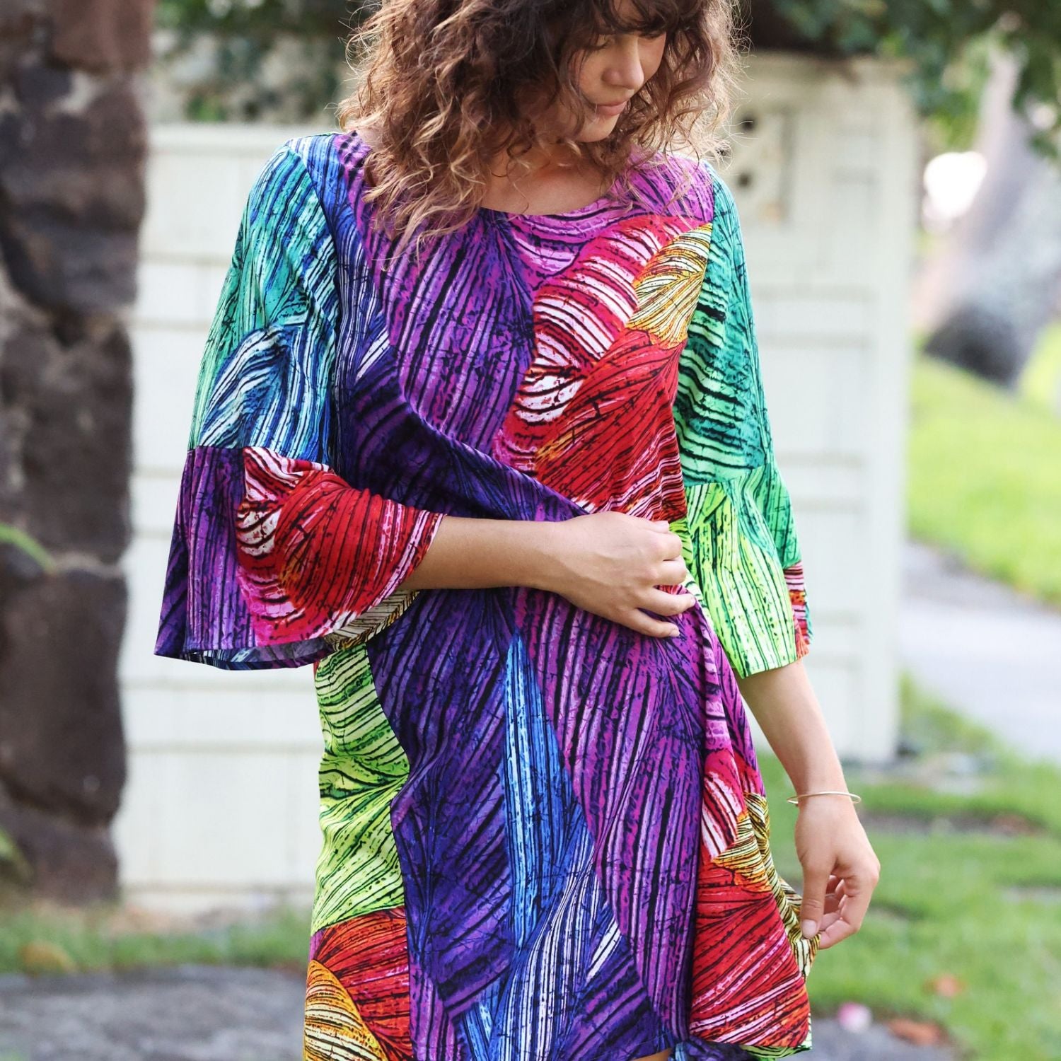 A woman wearing a colorful, patterned dress with three-quarter bell sleeves, standing outdoors in front of a light-colored wall and trees.