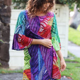 A woman wearing a colorful, patterned dress with three-quarter bell sleeves, standing outdoors in front of a light-colored wall and trees.