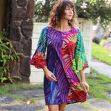 A woman wearing a vibrant, multicolored abstract-patterned dress with three-quarter bell sleeves, standing outdoors in a garden setting.
