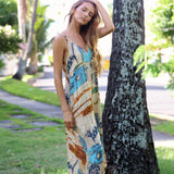 A woman wearing a sleeveless maxi dress with a colorful abstract print leans against a large tree trunk in a sunny, tropical outdoor setting.