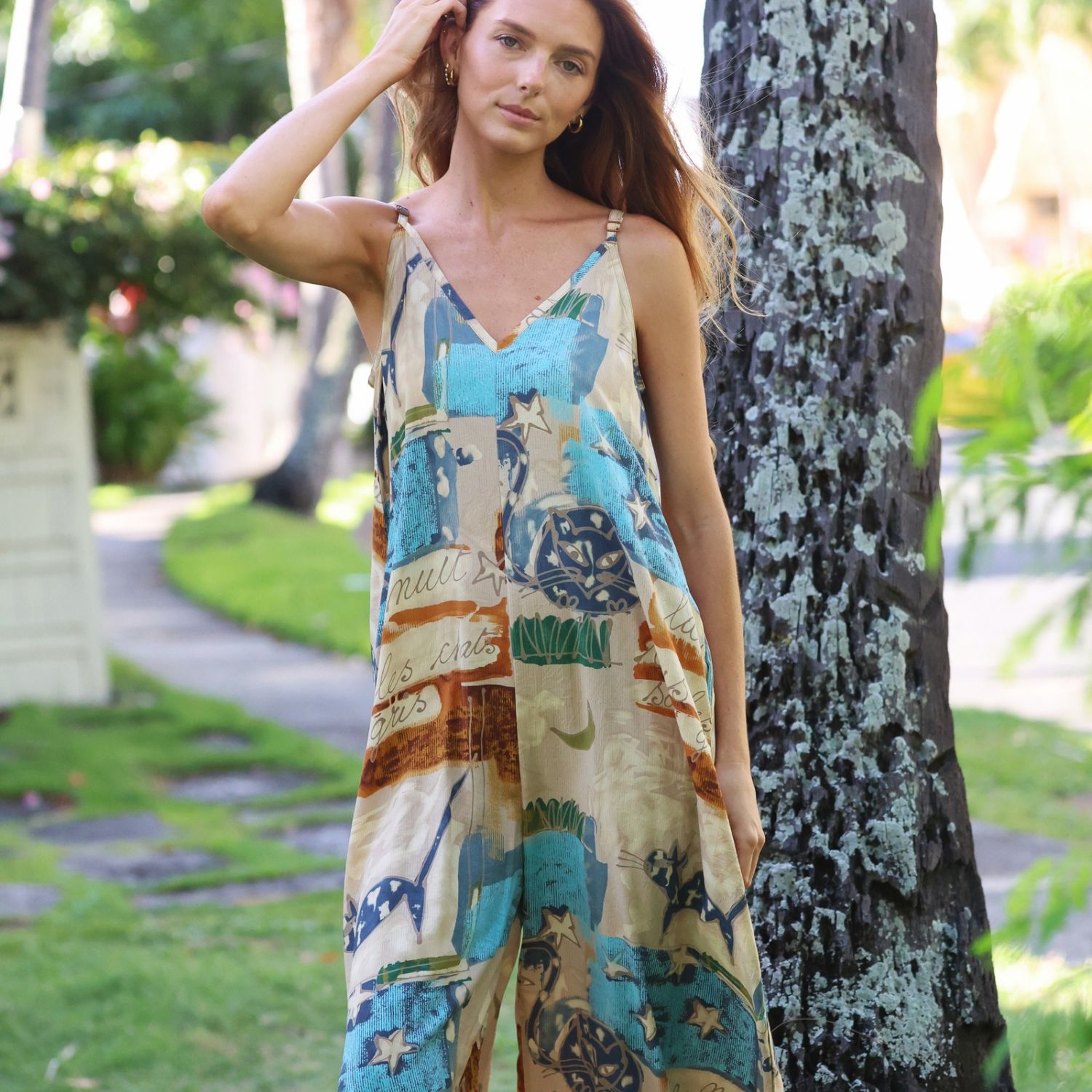 A woman wearing a sleeveless V-neck jumpsuit with a colorful abstract print in blue, beige, and brown, standing outdoors next to a tree trunk in a garden setting.