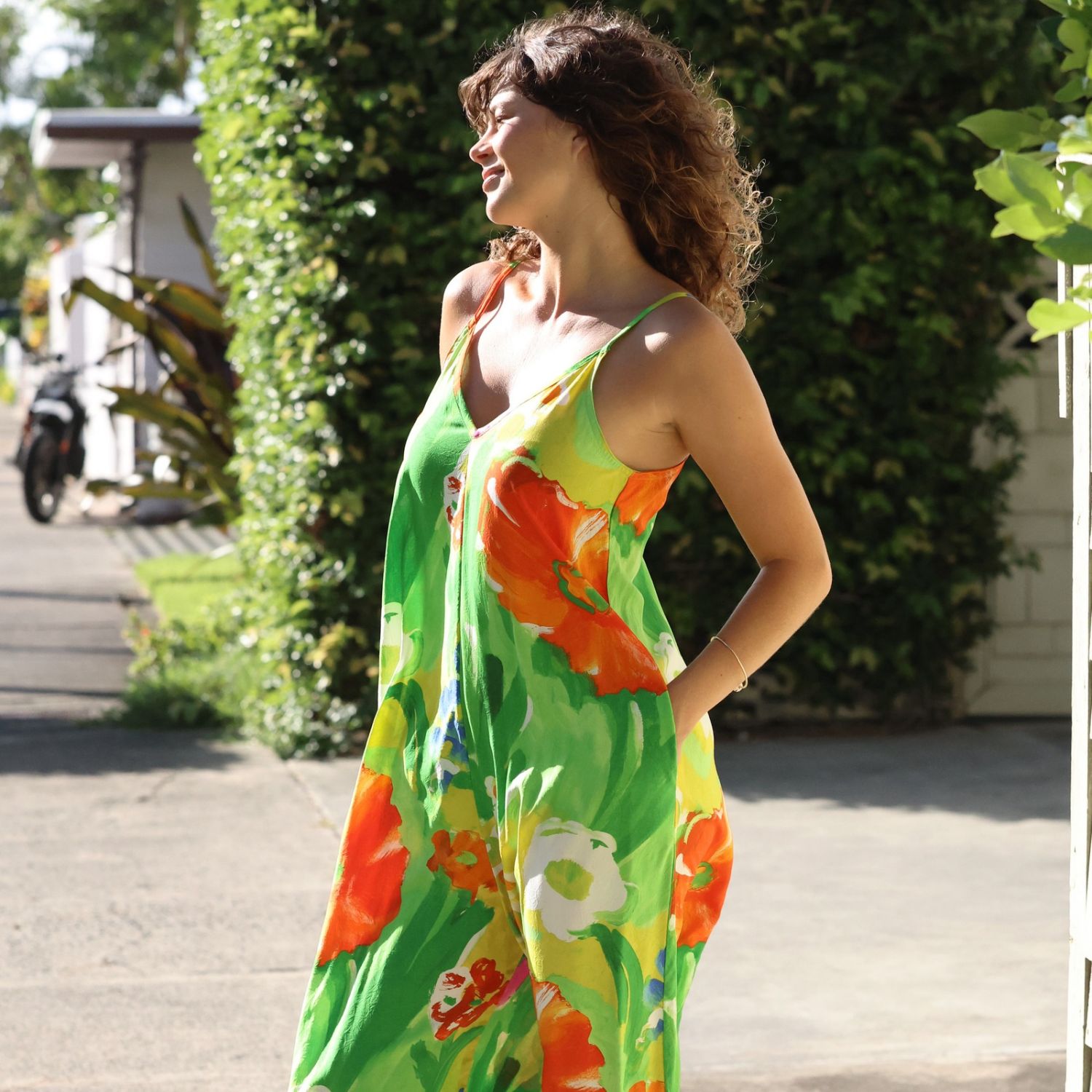 A woman wearing a vibrant green sleeveless maxi dress with a bold orange, yellow, and white floral print, standing outdoors on a paved path with green foliage in the background.
