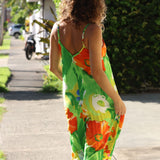 A woman viewed from behind wearing a bright green sleeveless jumpsuit with orange and yellow floral print, walking on a sunny outdoor sidewalk.