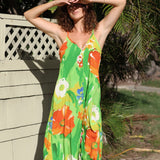 A woman wearing a vibrant green sleeveless maxi dress with a bold orange, white, and blue floral print, posing outdoors in front of a wooden fence and palm trees.