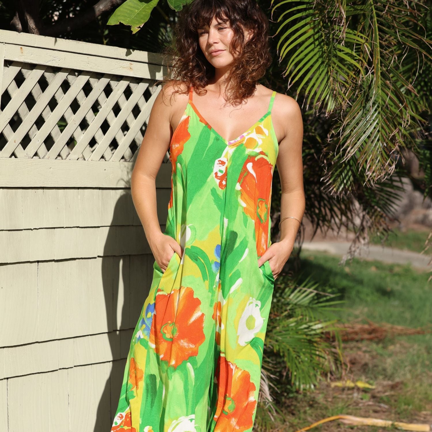 A woman wearing a vibrant green sleeveless maxi dress with a bold orange, yellow, and white floral print, standing outdoors next to a light-colored wooden fence with palm leaves in the background.