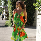 A woman wearing a vibrant green sleeveless maxi dress with a bold orange, yellow, and white floral print, standing outdoors on a paved path with green foliage and a white fence in the background.