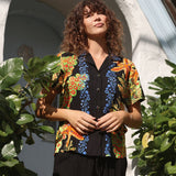 A woman wearing a short-sleeve button-up shirt with a vibrant multicolor floral and abstract print over a black base, standing outdoors in front of a white arched wall with green plants.