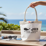White tote bag with JIB logo and text held by a hand on a table with ocean view