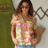 Model wearing colorful Niihau scenic print top with jeans shorts standing infront of a surfboard.