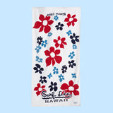 A full-length beach towel with a bold red, white, and blue hibiscus and flower pattern is displayed vertically against a light blue background. The white towel is covered with large red flowers, medium-sized dark navy blue flowers, and smaller light blue floral accents. The towel is oriented with the text “Since 1964” upside down near the top edge, and the logo “Surf Line HAWAII” in red script text appears near the bottom.
