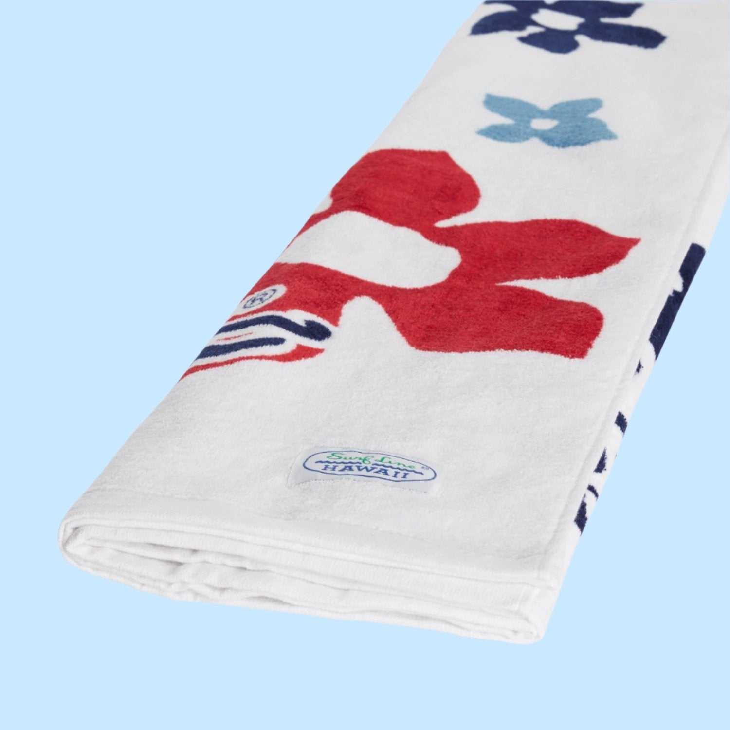 A close-up, angled view of the same white beach towel, which is folded or rolled. The pattern, featuring a large red flower, is partially visible. A light blue woven tag with the “Surf Line HAWAII” logo in white and blue text is sewn into the edge of the towel. The background is solid light blue.