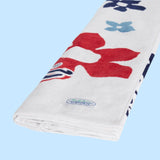 A close-up, angled view of the same white beach towel, which is folded or rolled. The pattern, featuring a large red flower, is partially visible. A light blue woven tag with the “Surf Line HAWAII” logo in white and blue text is sewn into the edge of the towel. The background is solid light blue.
