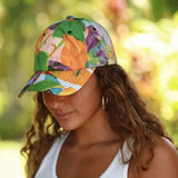 A woman wearing a colorful baseball cap with a vibrant floral and abstract print in green, orange, purple, and white, photographed outdoors with a soft, blurred green background.