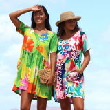 Two women wearing colorful floral mini dresses stand outdoors against a bright sky. The dress on the left is bright green with orange, pink, and blue floral print, and the dress on the right is white with a multicolor floral pattern.