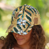 A woman wearing a baseball cap with a tropical floral pattern in beige, green, brown, and light blue, photographed outdoors with a blurred green background.