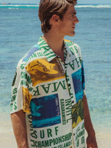A man wearing the white and multi-colored surf-themed aloha shirt and light blue shorts stands on a sandy beach, looking to the side with the clear, shallow ocean water in the background.