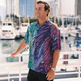 Man wearing a colorful patterned shirt standing by a marina with boats and buildings in the background.