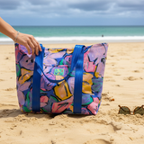 一只手拿着一个色彩鲜艳、印有抽象图案的帆布包，站在沙滩上。
