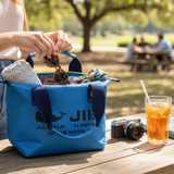 A lifestyle photo of a medium sized, vibrant blue JIB tote bag on a wooden picnic table. A person is reaching into the bag, which is packed with a patterned towel, sunglasses, and a colorful shirt. Next to the bag sits a silver film camera and a glass of iced tea with lemon. The background is a soft focus sunny park.