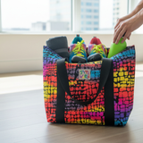 Colorful tote bag with a pattern filled with shoes and a green bottle, placed on a wooden floor.