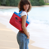 A woman stands on a beach with her back to the camera, looking over her shoulder while wearing a red JIB tote with a bright blue top border and blue handles.