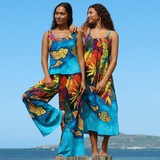 Two women in colorful outfits standing on a wooden platform with a clear blue sky and ocean background.