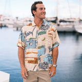 Man wearing a colorful patterned shirt standing by a waterfront with boats in the background