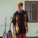 Man wearing a colorful floral shirt standing outdoors with paddle sports equipment in the background.