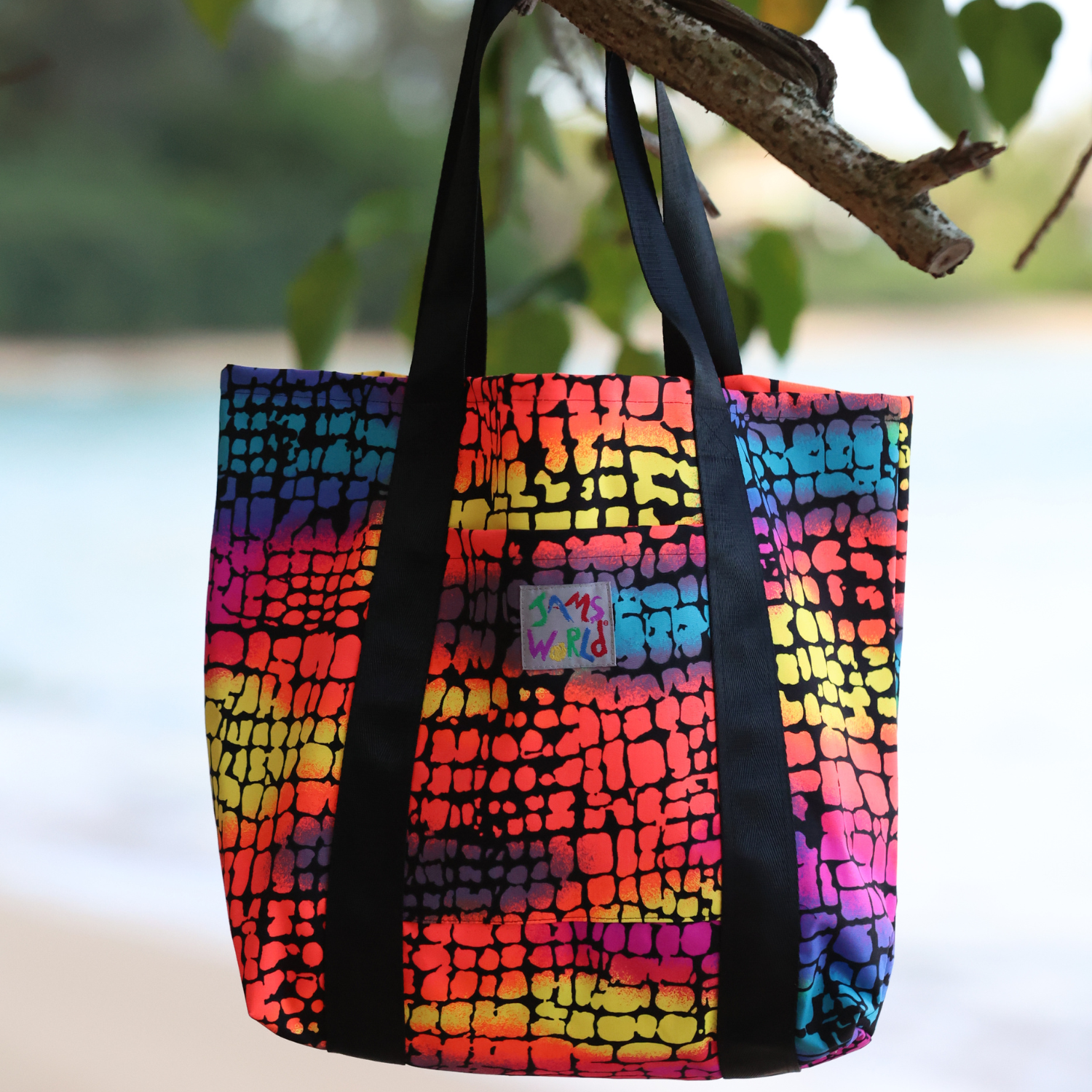 A vibrant, multicolored “Jams World” tote bag with a rainbow crocodile-skin pattern, hanging from a tree branch against a blurred beach background.