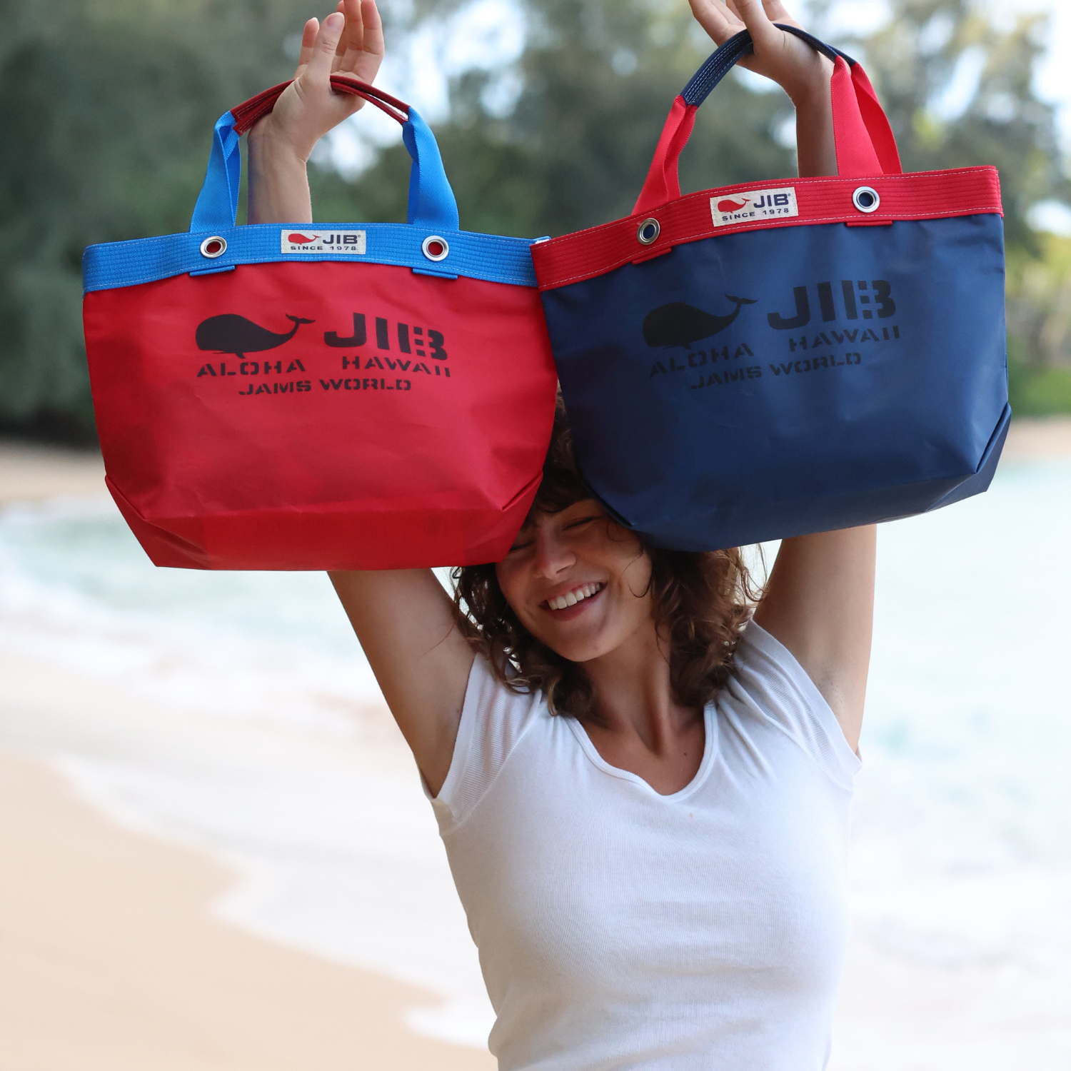 Sur une plage, une femme souriante brandit deux sacs fourre-tout JIB : l'un rouge avec des bordures et des anses bleues, et l'autre bleu marine avec des bordures et des anses rouges.