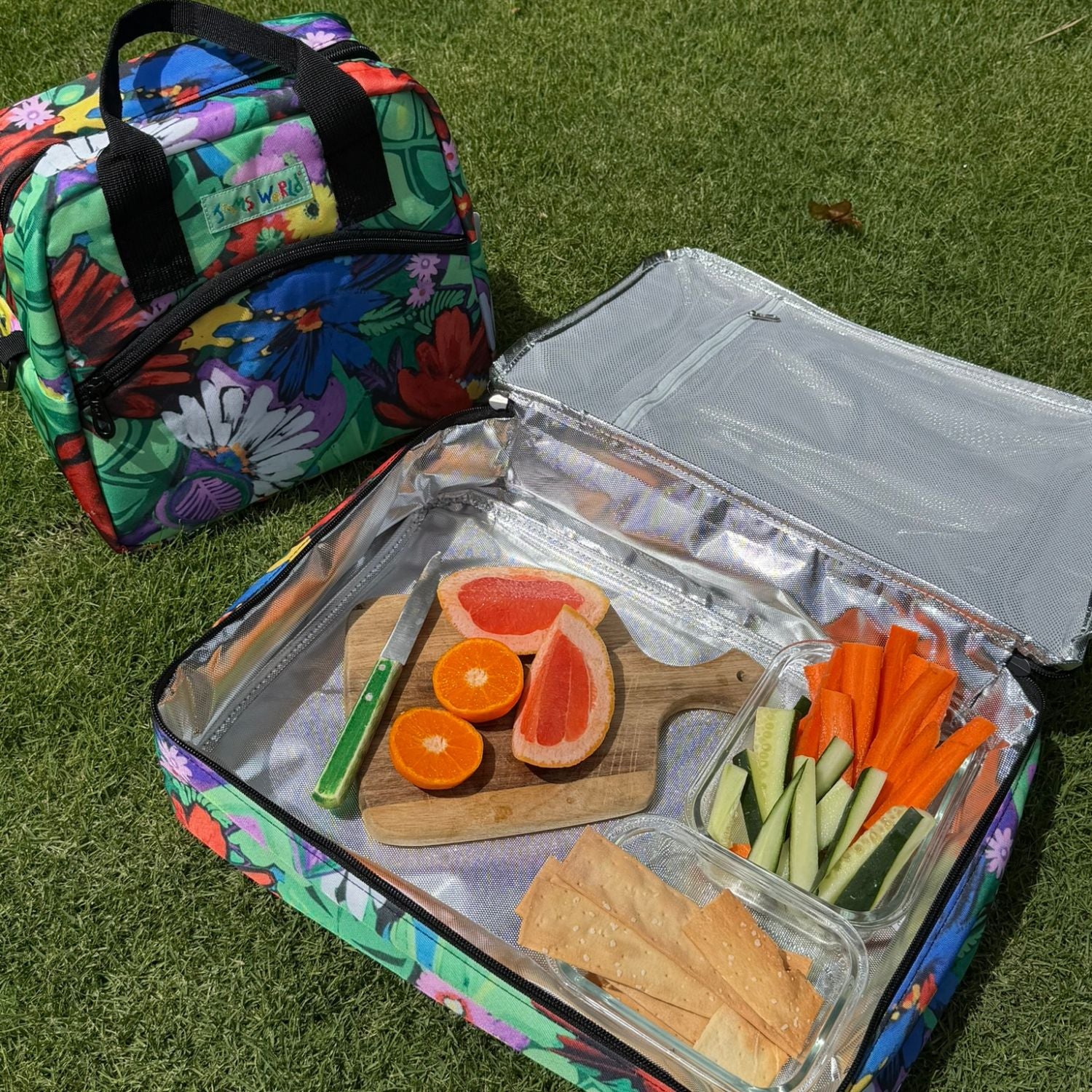 Bolsa isotérmica de colores con una tabla de cortar de madera que contiene frutas y verduras en rodajas sobre césped.