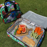 Colorful insulated lunch bag with a wooden cutting board containing sliced fruits and vegetables on grass.