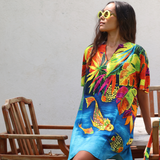 A woman wearing a vibrant tropical print tunic dress with colorful fish and leaf patterns, styled with yellow sunglasses, sitting on a wooden chair against a light wall.