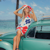 Woman in a colorful dress and hat sitting on a vintage teal car by the ocean.