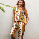 A woman wearing a matching short-sleeve button-up shirt and wide-leg pants set with a colorful tropical floral and bird print, posing against a textured white wall.