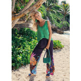 Mujer con blusa verde y pantalones coloridos caminando por una playa con vegetación.