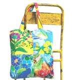 Un bolso de tela con asas estampadas multicolores, colgado de una silla o escalera de metal amarillo brillante y desgastado. El bolso presenta un mosaico de cuatro cuadrados con estampado floral vibrante en colores como amarillo, azul, rojo, morado y verde. El fondo es blanco puro.